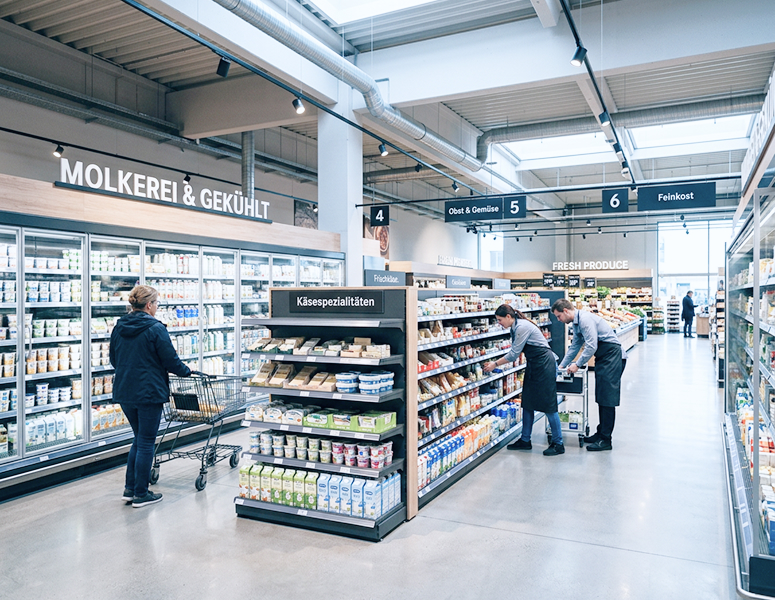 Moderner Supermarkt mit Kundin und Mitarbeitern zwischen Kühlregalen und Warenbereichen.